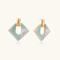 Geometric square enamel earrings with marbled turquoise and lavender pattern, gold U-shaped connector on cream background.